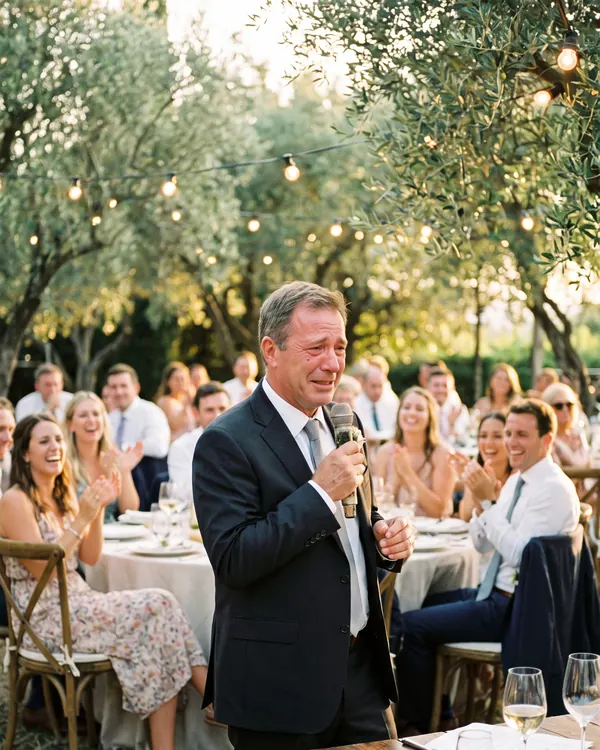 The father of the bride, eyes wet, gives a toast into a microphone while guests smile at the tables under olive trees.