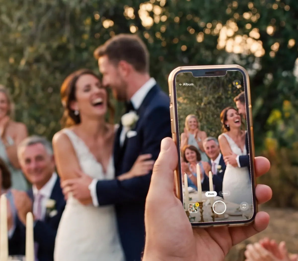 A guest films the couple's embrace on his phone, the Sharecam camera open on the screen, while the table applauds under the olive trees.